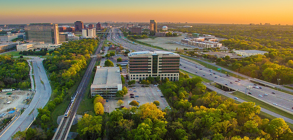Aerial view of Richardson, Texas business corridor at sunset with office buildings, Central Expressway (US-75), DART light rail, and lush green landscape.